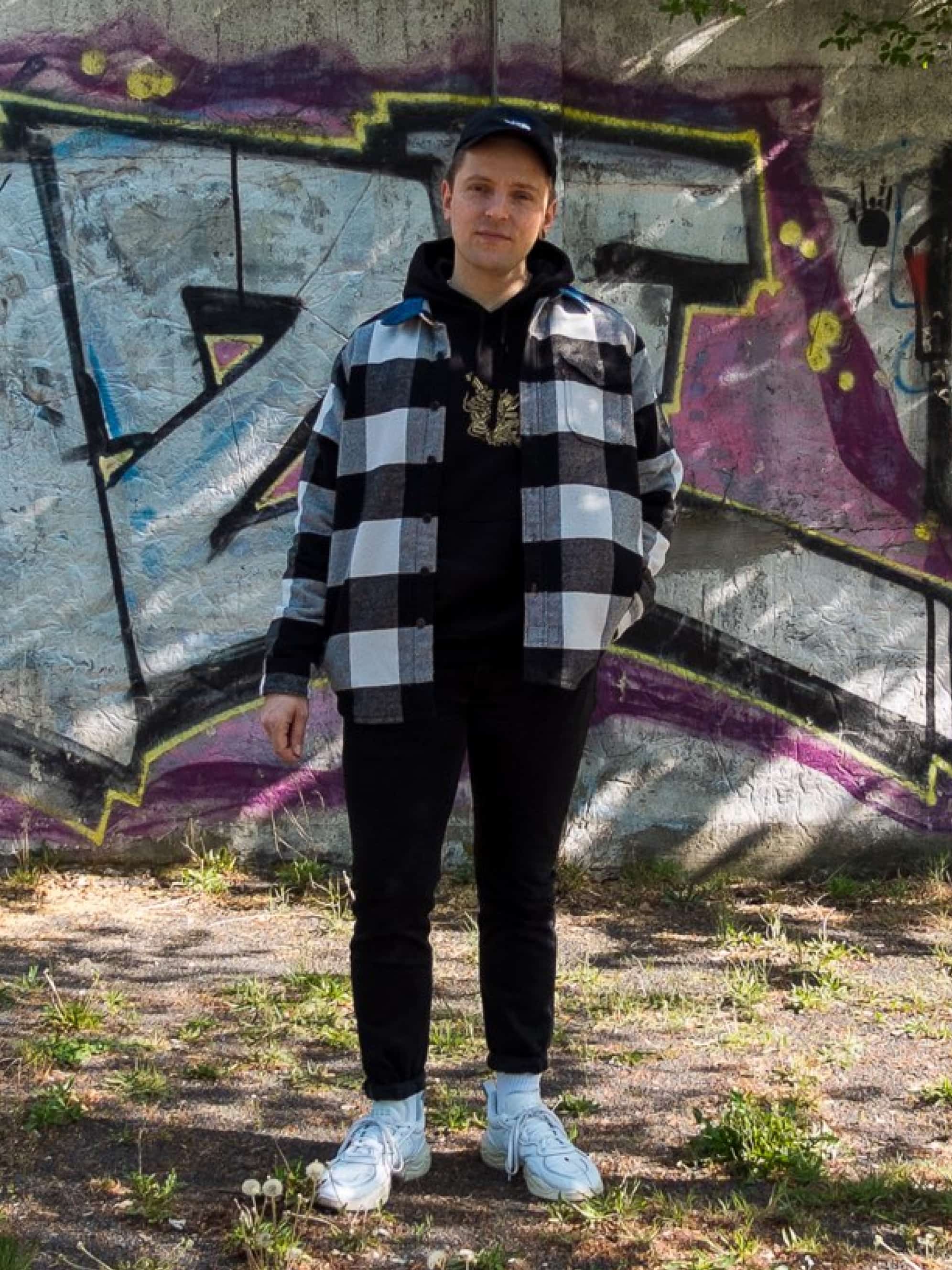 A man in a plaid shirt and black pants stands in front of graffiti.