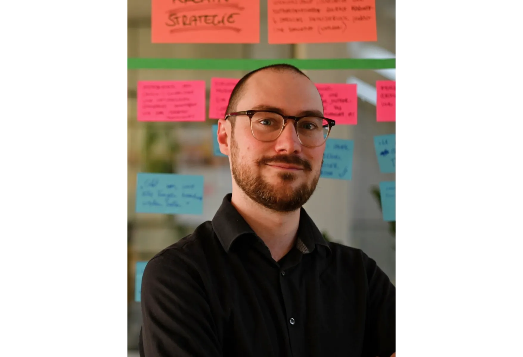 Person mit Brille und Bart steht mit verschränkten Armen vor einer Glaswand mit bunten Haftnotizen.