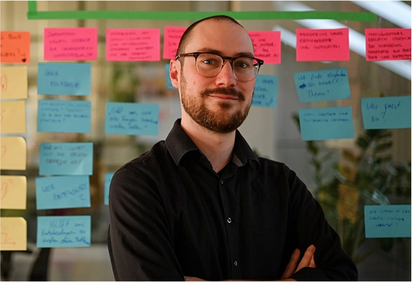 Person mit Brille und Bart steht mit verschränkten Armen vor einer Glaswand mit bunten Haftnotizen.