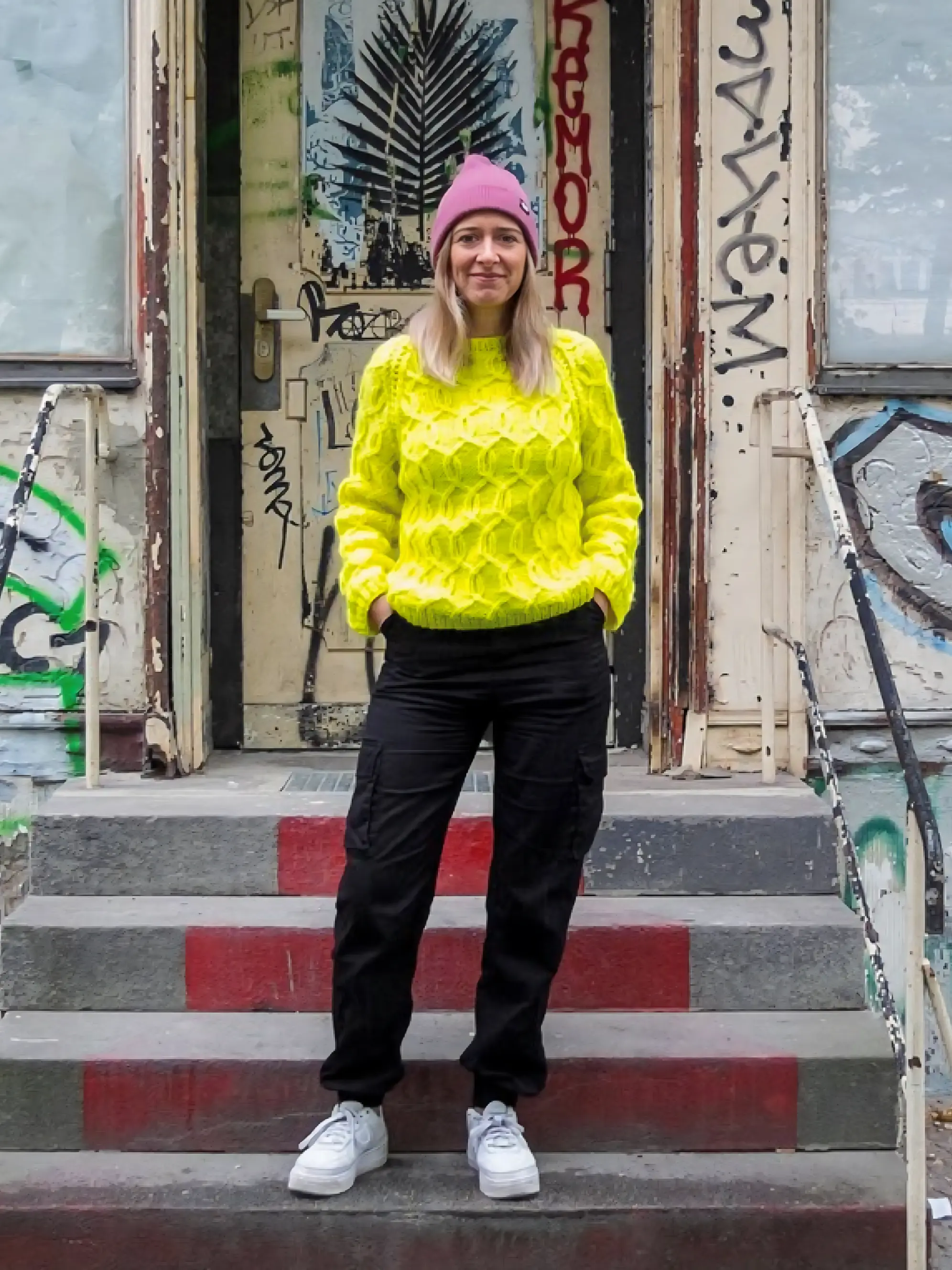 Junge Frau mit gelbem Strickpullover und pinker Mütze vor Hauseingang