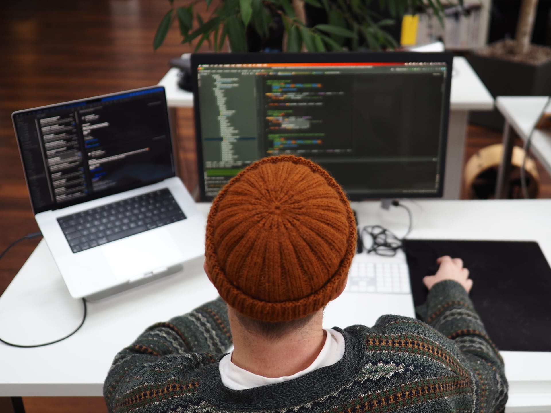 Ein Mann mit Hut sitzt an einem Schreibtisch mit einem Laptop und einem Bildschirm.