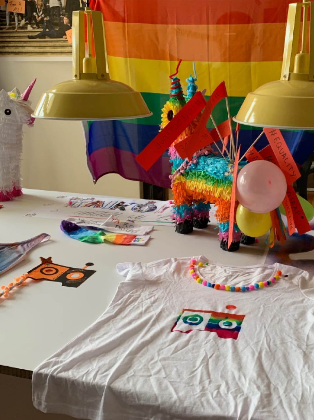A table with an LGBTQ+ rainbow flag, balloons, a colorful pinata and colorful painted shirts.