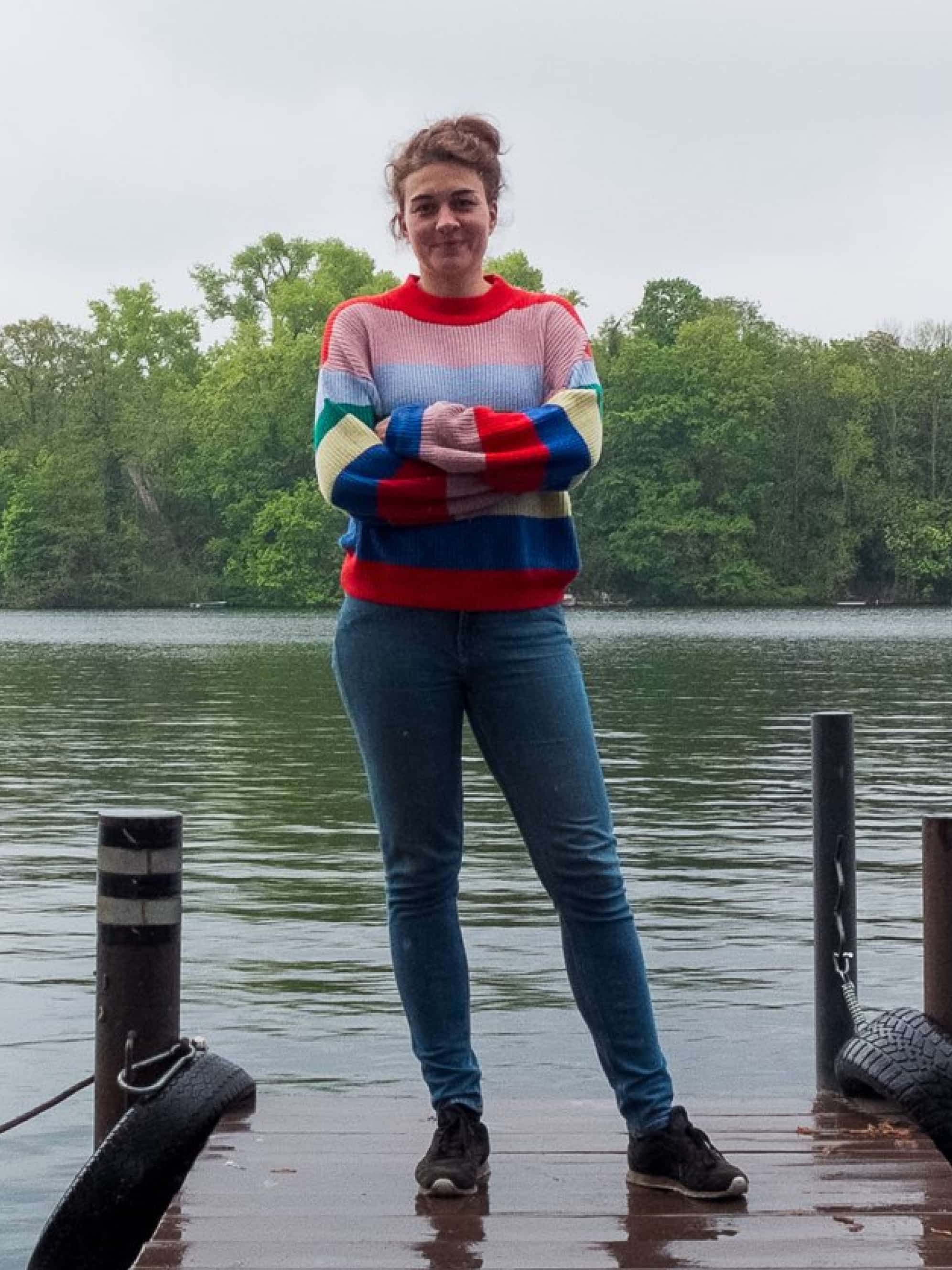 A woman in a colorful knitted sweater stands on a jetty in front of Lake Tegel.