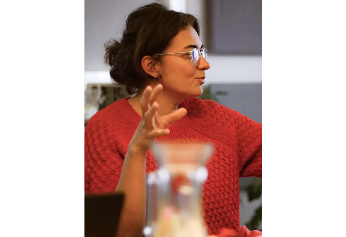 Person mit Brille und rotem Strickpullover spricht und gestikuliert lebhaft, seitlich aufgenommen in einem Raum mit entspannter Atmosphäre.