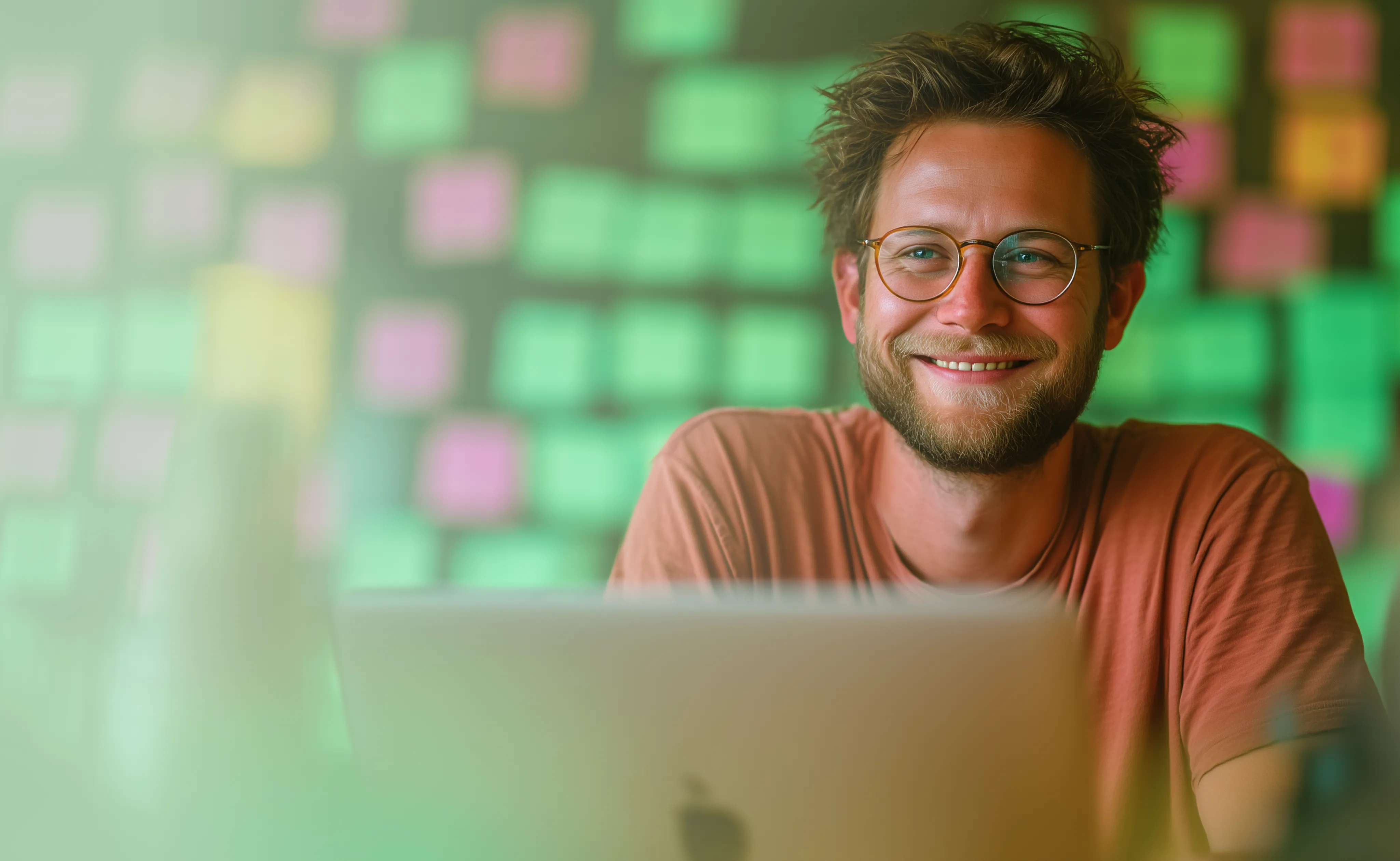 Lächelnde Person mit Brille und Bart sitzt vor einem Laptop, im Hintergrund eine Wand voller bunter Haftnotizen.