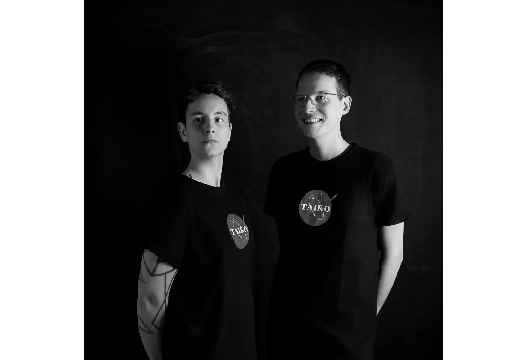 Zwei Personen in schwarzen T-Shirts schauen stehen vor schwarzem Hintergrund (s/w)