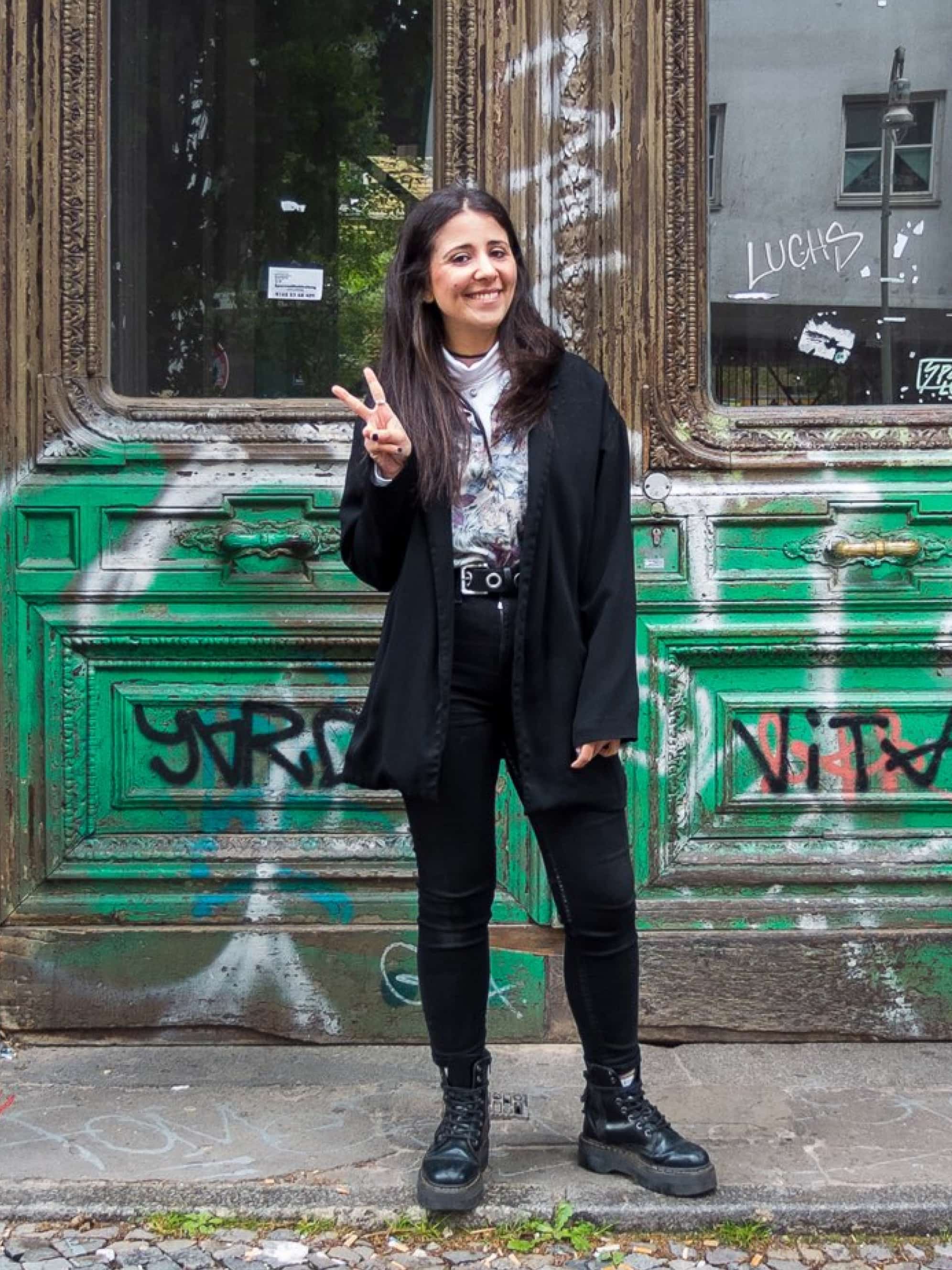 A woman in a black jacket and jeans stands in front of a green door and makes a peace sign with her right hand.