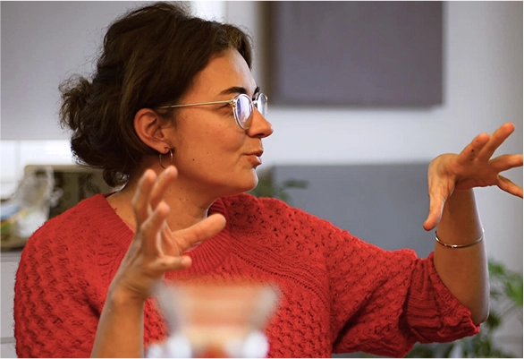 Person mit Brille und rotem Strickpullover spricht und gestikuliert lebhaft, seitlich aufgenommen in einem Raum mit entspannter Atmosphäre.