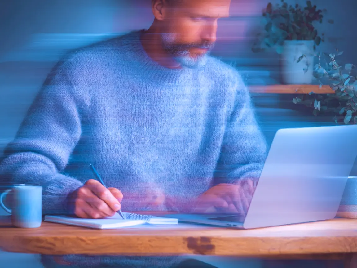 Person mit blauem Pullover arbeitet an einem Laptop und macht sich Notizen in ein Notizbuch, dynamischer Effekt durch Bewegungsunschärfe.