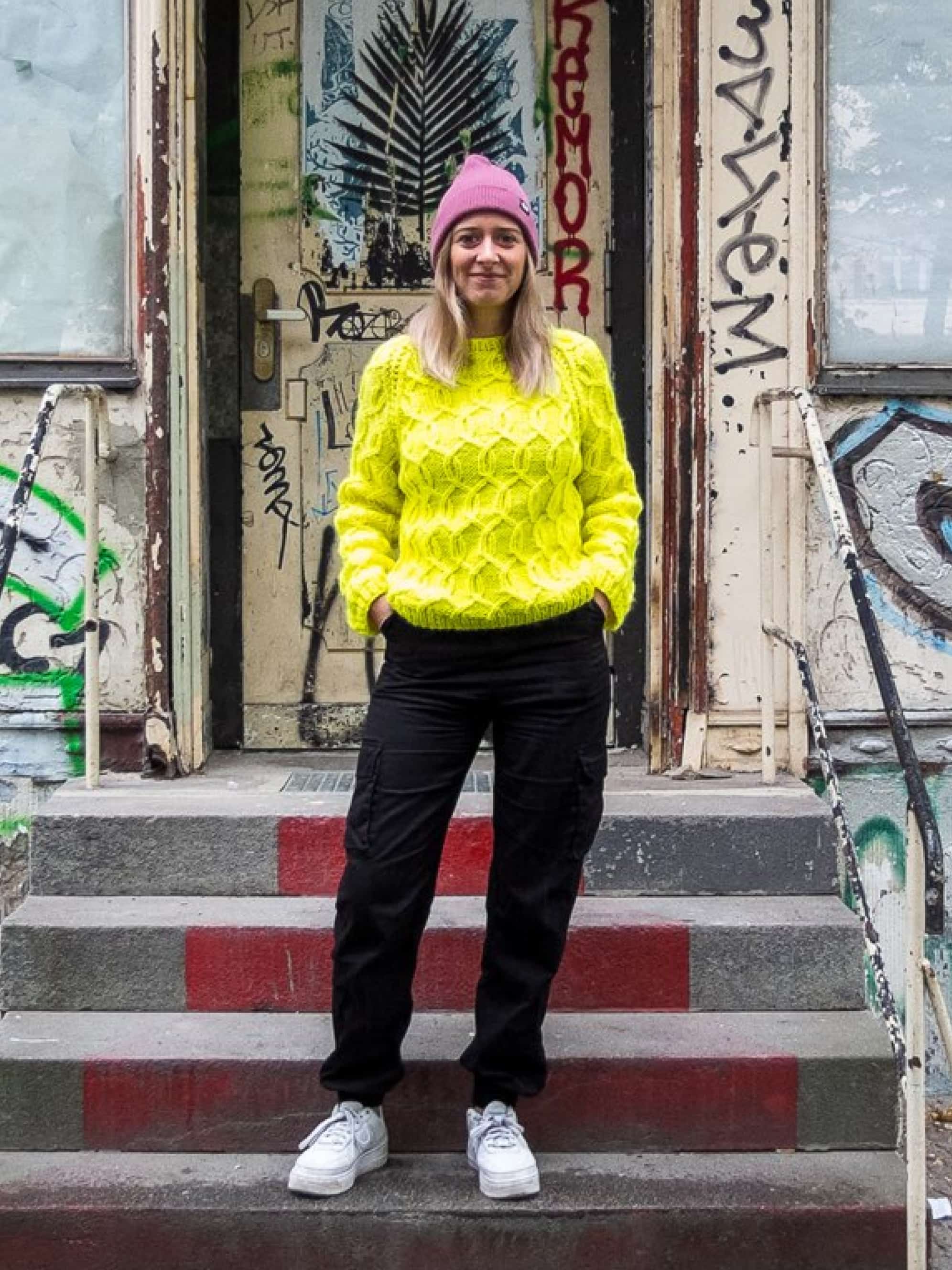 A woman in a yellow sweater and black trousers with a pink cap is standing on a staircase.