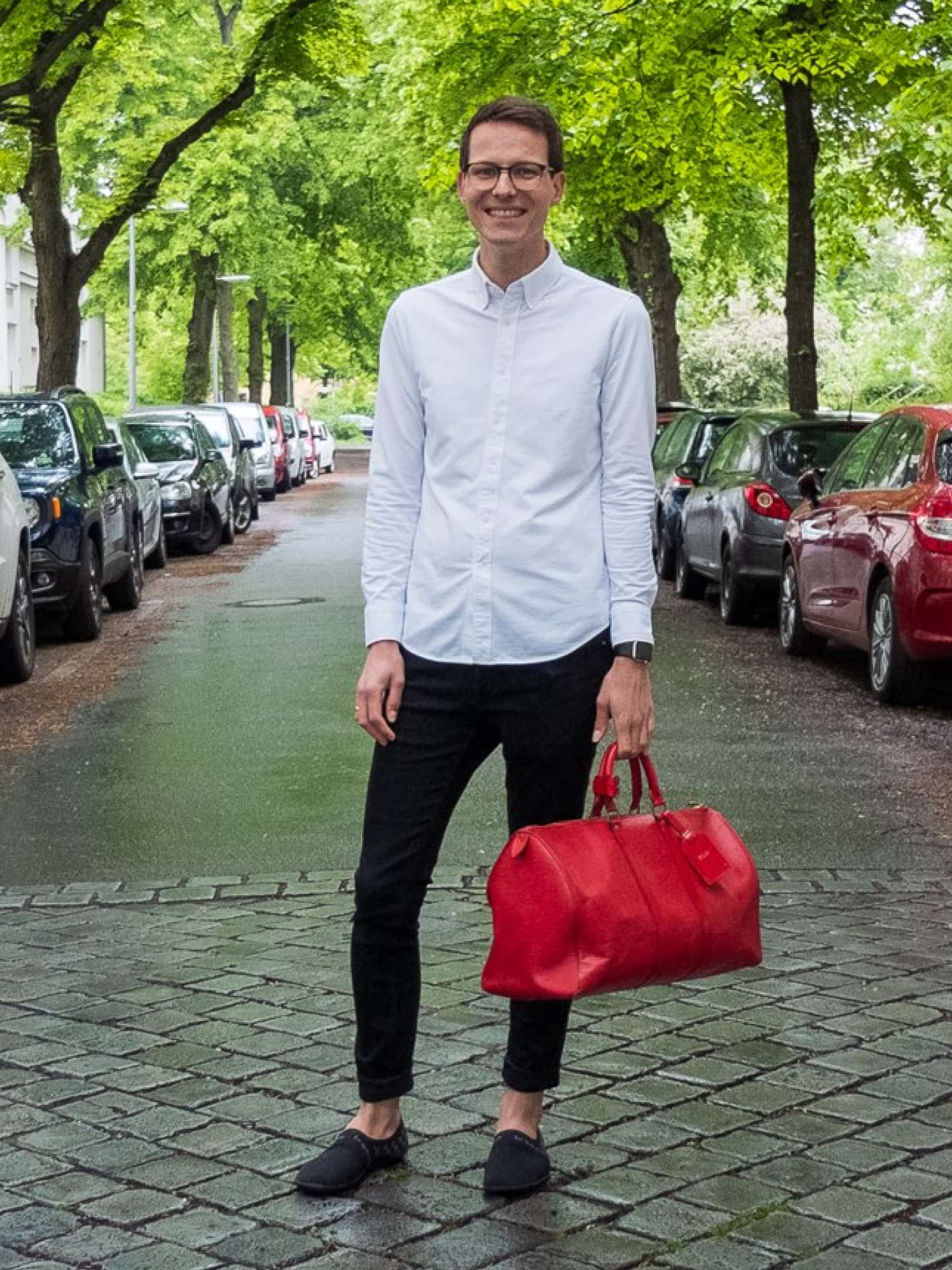 A man in a white shirt and black trousers holds a red bag.