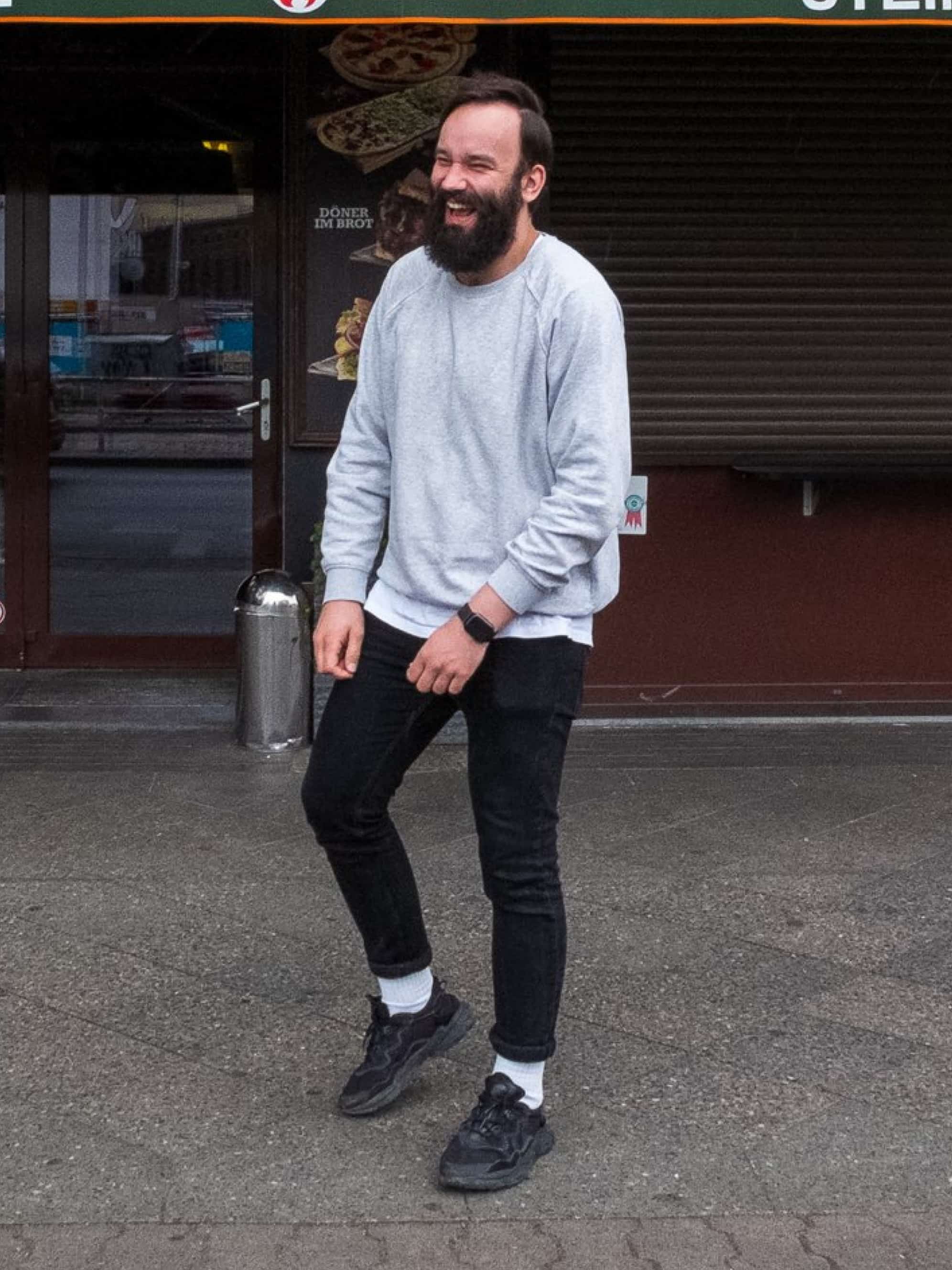 A man in a gray sweater stands on the sidewalk in front of a store and laughs.
