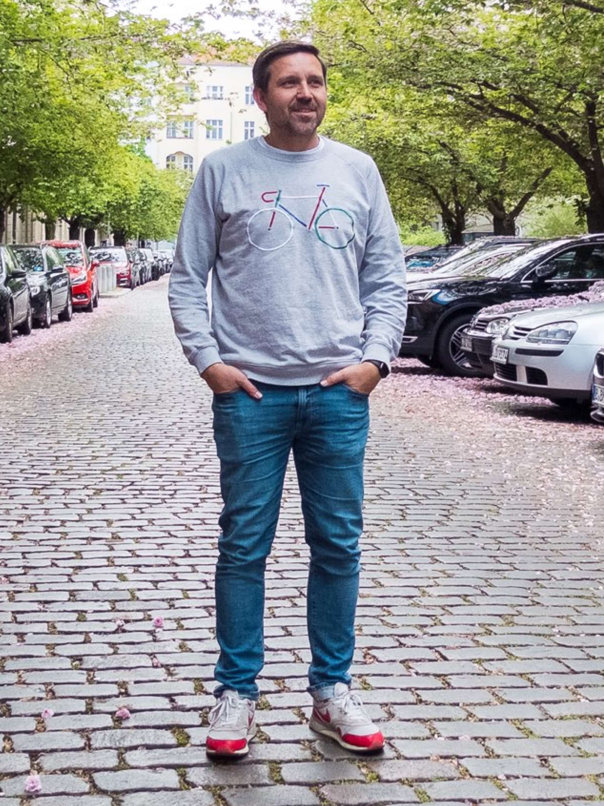A man in a gray sweatshirt and jeans stands on a cobblestone street.