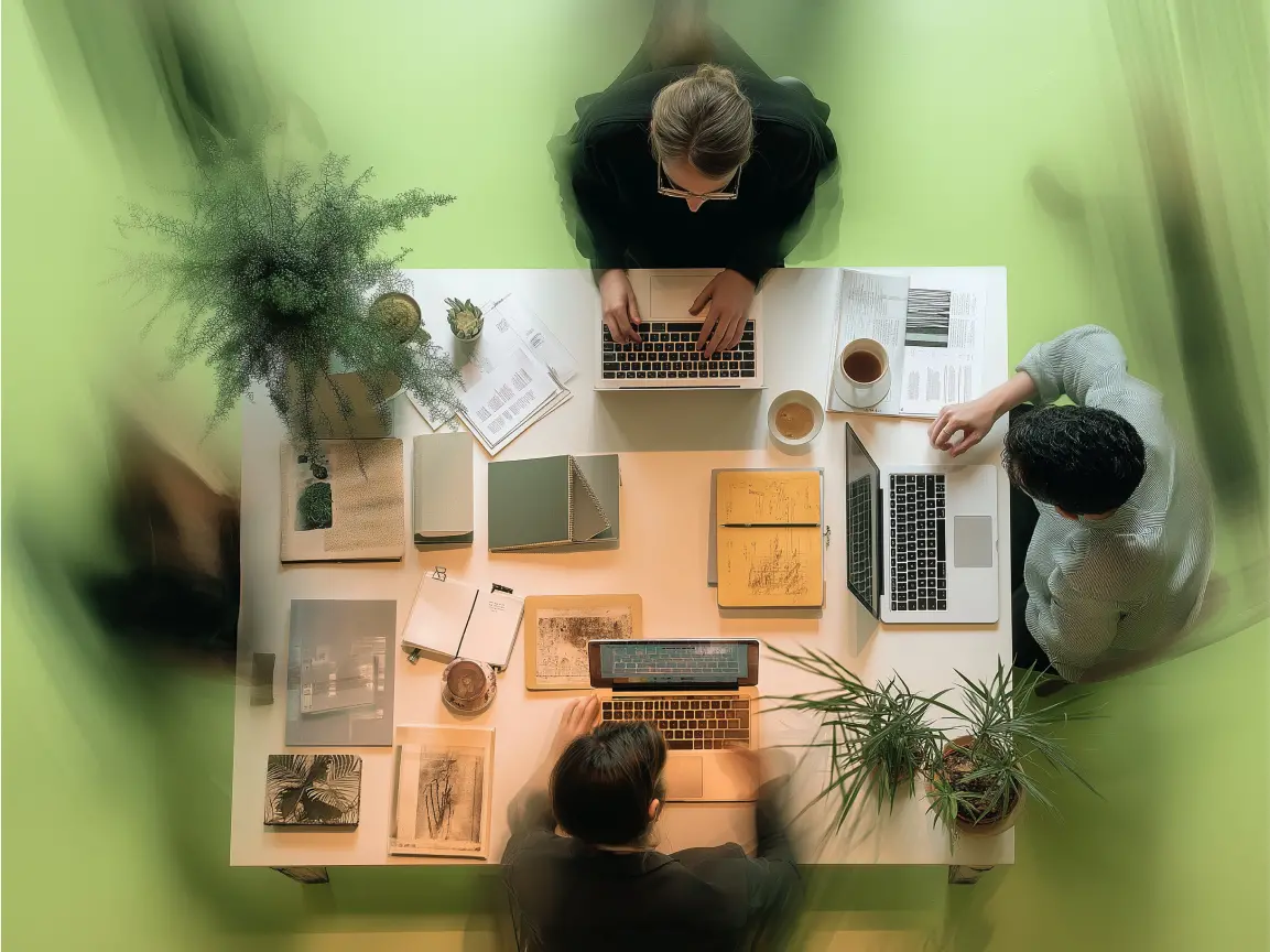 Vier Personen arbeiten gemeinsam an einem Tisch mit Laptops, Notizbüchern und Pflanzen, von oben fotografiert in einer modernen Arbeitsumgebung.
