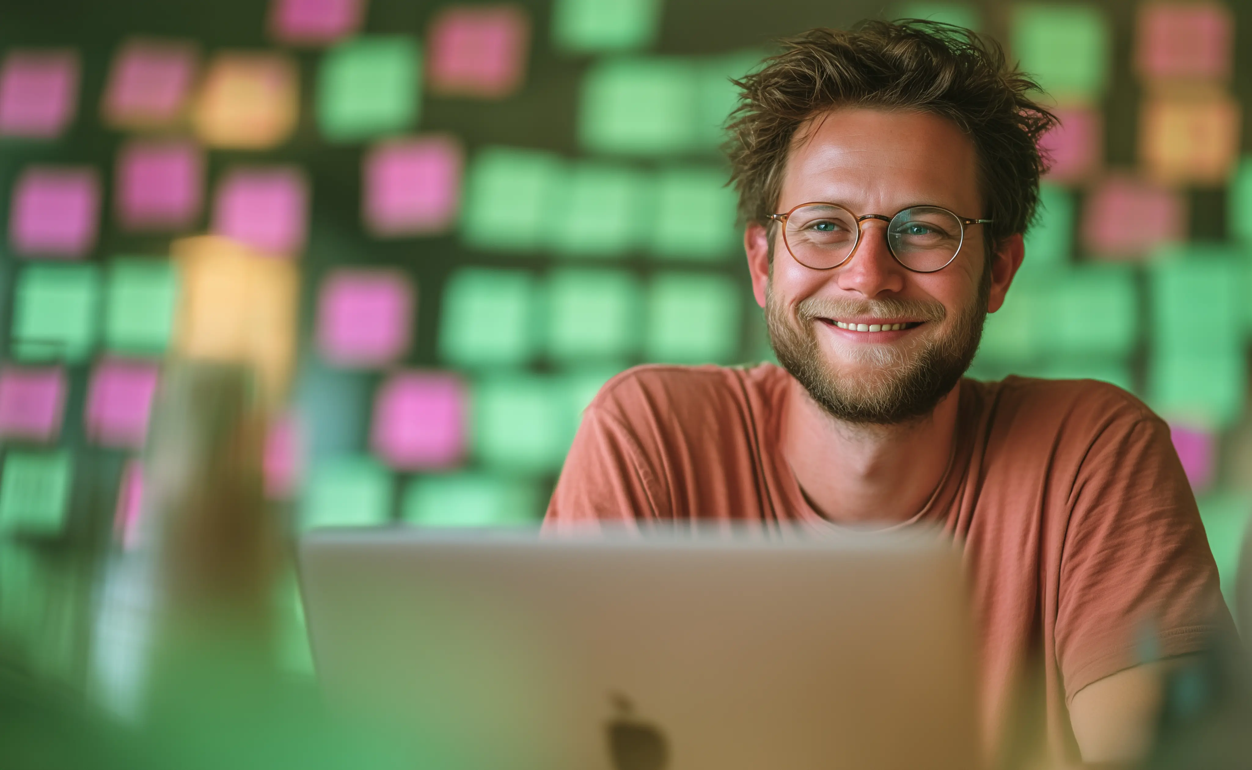 Mann sitzt vor mit Post-It-Zetteln beklebter Wand und lächelt über seinen Laptop hinweg in die Kamera