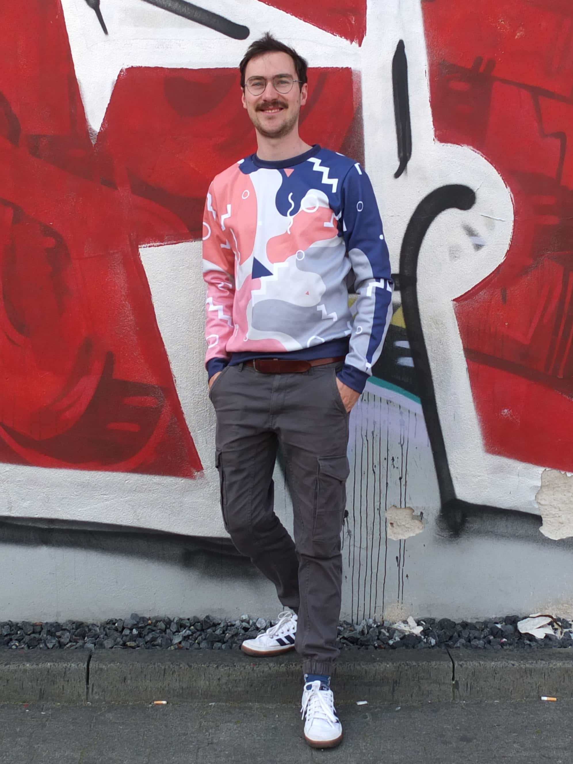 A man in a colorful sweater leans against a graffiti wall.