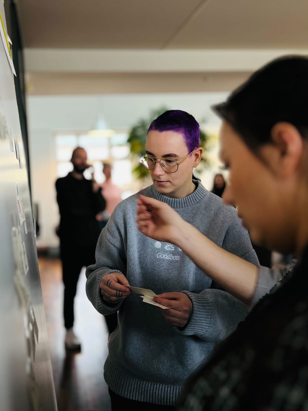 A person with short purple hair stands in front of a whiteboard with sticky notes in hand.